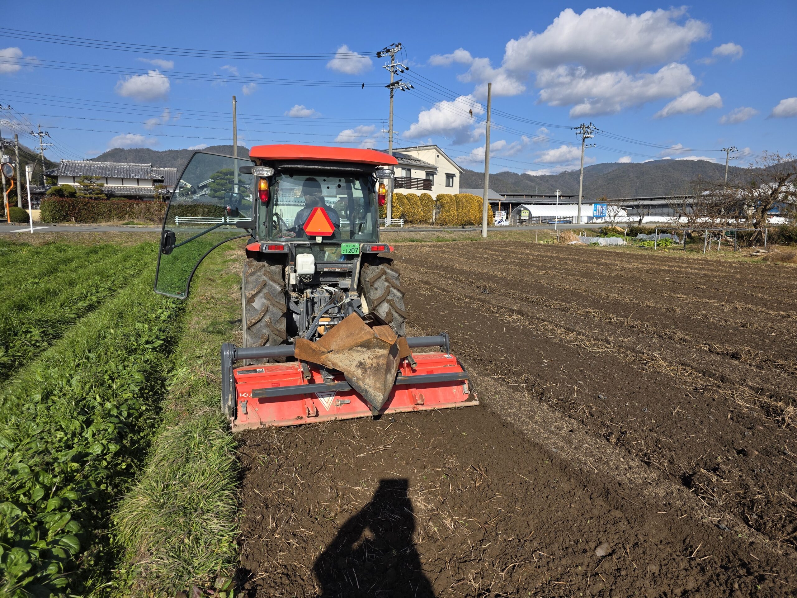 亀岡の青空の下、広大な農地で赤いトラクターを運転し耕耘作業を行う様子